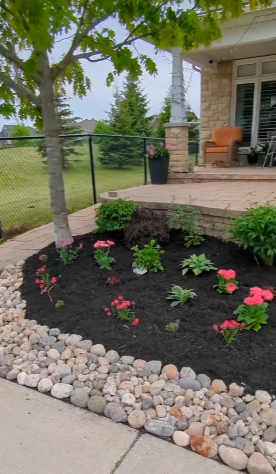 Front entrance garden bed featuring crisp river rock border, black mulch, dianthus flowers, and hostas